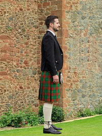 Man wearing a traditional Scottish kilt standing against a stone wall.