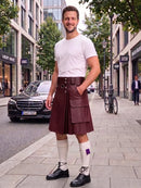 A man stands on a city sidewalk wearing a dark burgundy leather kilt with a white t-shirt, white knee-high socks, and black leather shoes. A Mercedes-Benz car and a bicycle are visible in the background of the urban street scene.