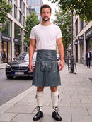 A man stands on a city sidewalk wearing a white crew-neck t-shirt and a dark gray faux-leather kilt with front pockets and a drawstring waist. He is also wearing white knee-high socks and black leather shoes.