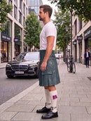 A man standing on a city sidewalk wearing a dark green kilt, white t-shirt, white socks, and black shoes, with a street scene and parked cars in the background.