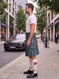 A man standing on a city sidewalk wearing a dark green kilt, white t-shirt, white socks, and black shoes, with a street scene and parked cars in the background.
