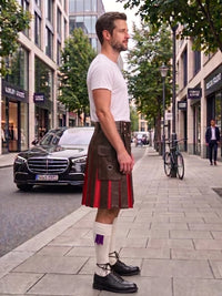 A man standing on a city sidewalk wearing a brown leather kilt with red pleated fabric, a white t-shirt, white socks, and black leather shoes. A black Mercedes-Benz car and a bicycle are parked nearby on the street.