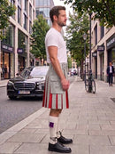 A man standing on a city sidewalk wearing a light gray kilt with red and white pleats, a white t-shirt, white knee-high socks, and black leather shoes. A black Mercedes-Benz car and a bicycle are parked on the street behind him.