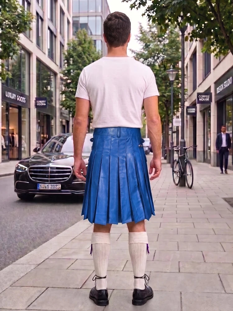 A man standing on a city sidewalk, viewed from behind, wearing a white t-shirt, a bright blue pleated leather kilt, white knee-high socks, and black shoes. Buildings, trees, a black Mercedes, and a bicycle are visible in the background.