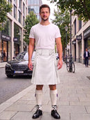 A man stands on a city sidewalk wearing a white short-sleeve t-shirt and a white kilt with belt loops, paired with white knee-high socks and black leather shoes. Buildings, trees, a parked black car, and a bicycle are visible in the background.