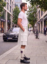 A man wearing a white short-sleeve t-shirt and a white pleated kilt with a white belt, standing on a city sidewalk next to a street with a black Mercedes-Benz car and trees.