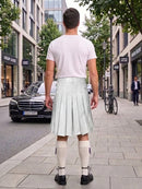 A man standing on a city sidewalk, viewed from behind, wearing a white short-sleeve T-shirt and a pale mint green pleated kilt with white knee-high socks and black shoes. Storefronts, trees, and a parked bicycle are visible in the background.