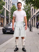 A man stands on a city sidewalk wearing a white t-shirt, a light grayish-green kilt with a silver-toned leather strap and pouch, cream knee-high socks, and black leather shoes. Modern buildings, trees, and a black Mercedes-Benz are visible in the background.